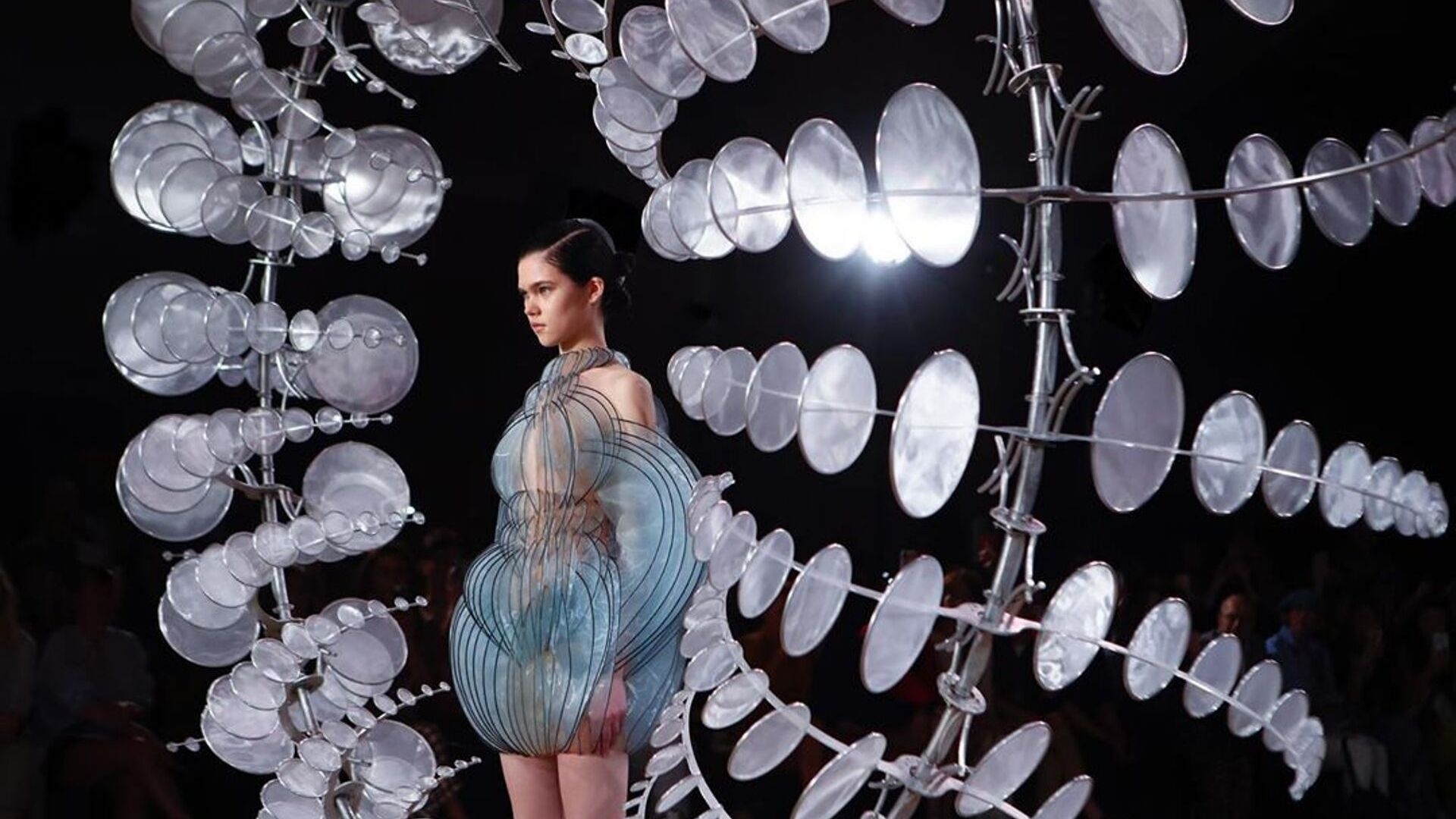 A model wears a futuristic dress surrounded by abstract art on a runway, highlighting digital influencer fashion.