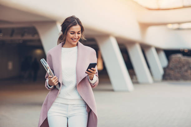 A stylish woman in a pink coat and white outfit looks at her phone while walking outdoors.